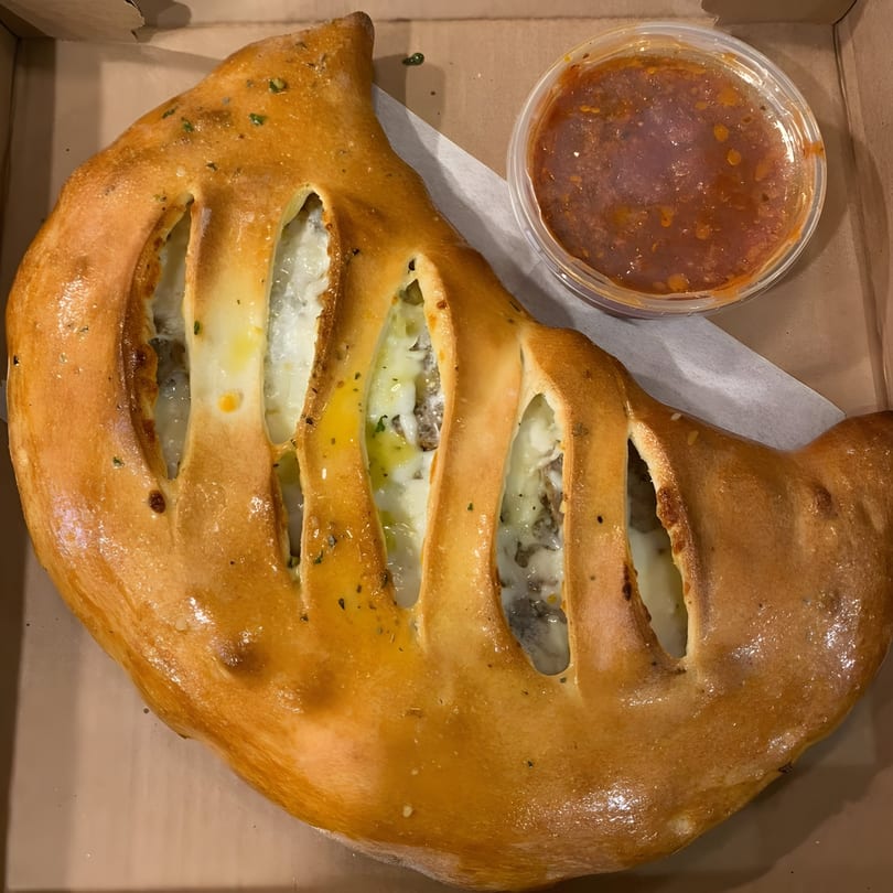 A golden-brown calzone with visible cheese and meat filling, served with a side of marinara sauce.