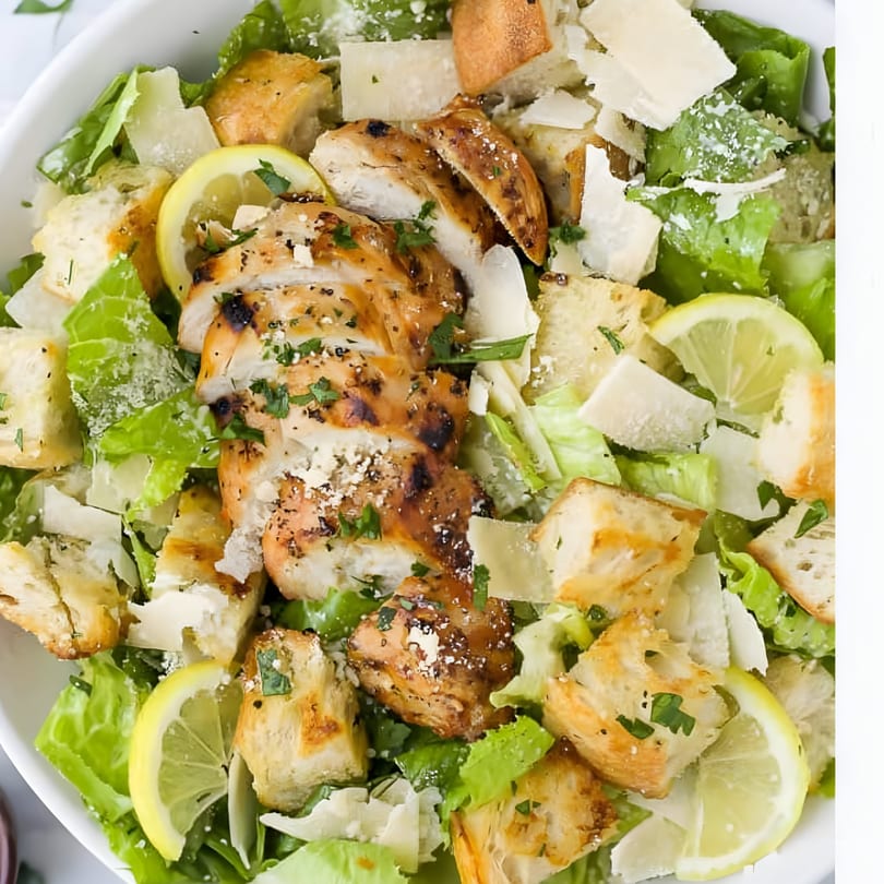 A close-up of a Caesar salad with grilled chicken, croutons, parmesan cheese, and lemon slices.