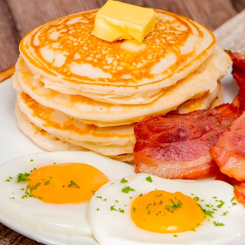 A diner breakfast with a stack of pancakes, butter, bacon, and two fried eggs.