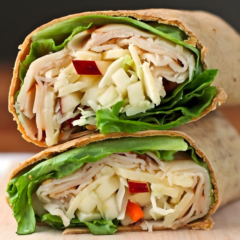Two halves of a turkey and apple wrap, filled with lettuce, shredded cheese, and red apples.